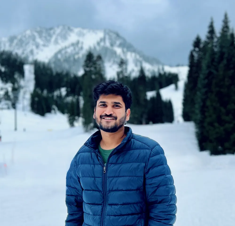 Abhi smiling in a blue puffer jacket in front of snowy mountains and pine trees